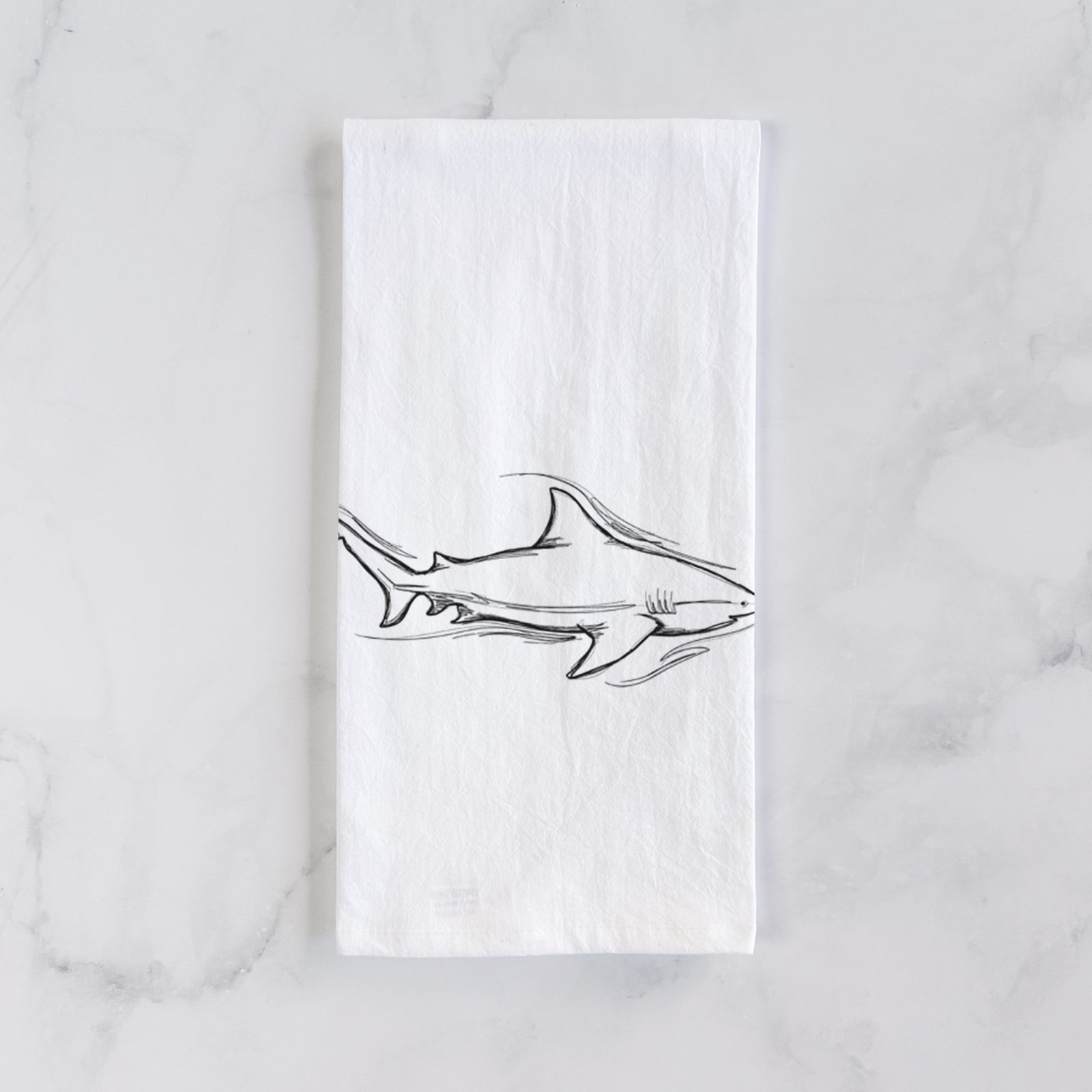 White cotton tea towel printed with a black ink illustration of a bull shark swimming, on a marble counter beside a round wooden cutting board and greenery.
