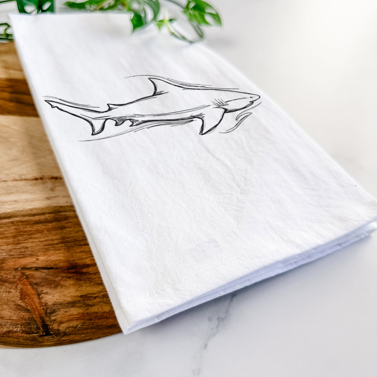 Close-up of a folded white tea towel on a wooden board, showing a black ink illustration of a bull shark swimming.