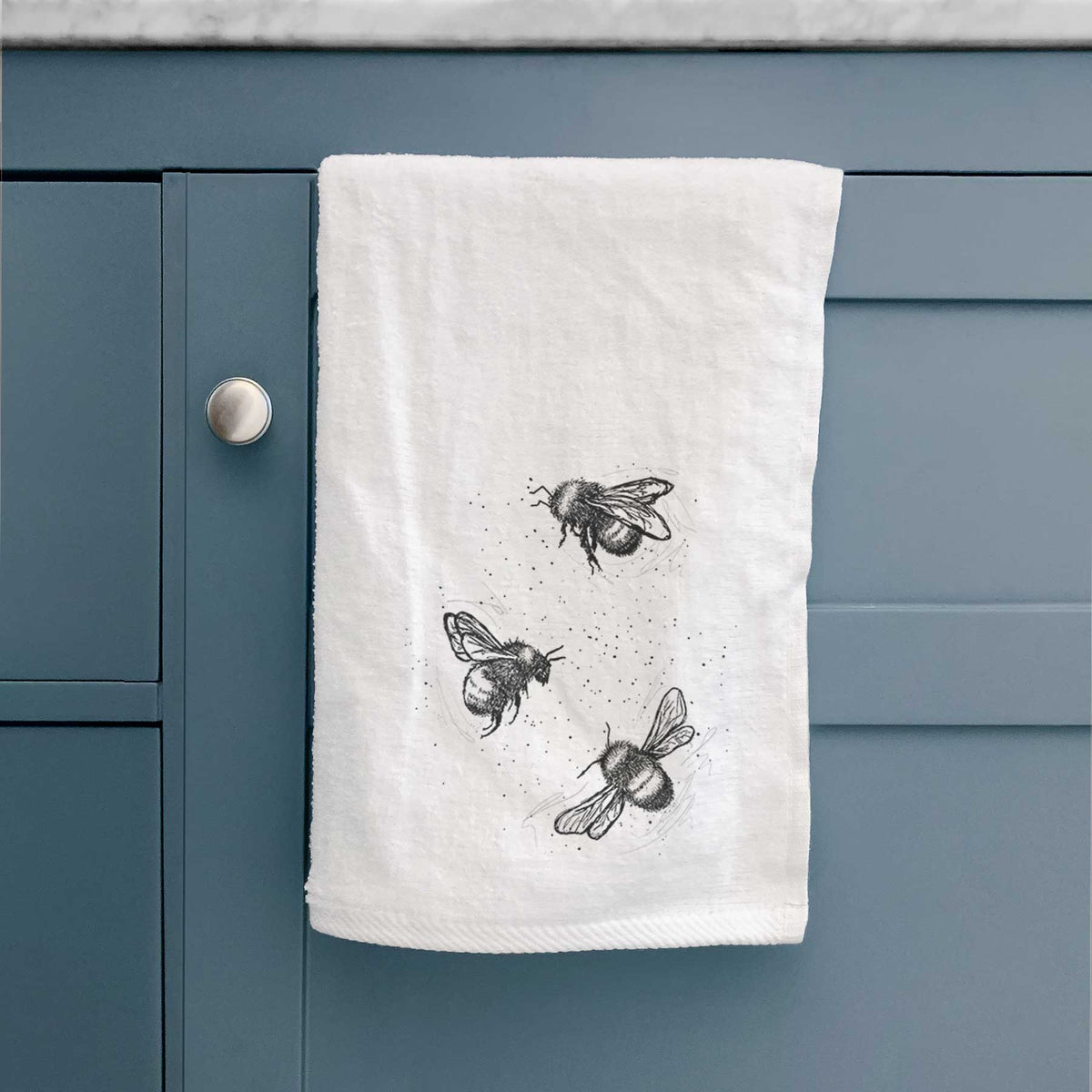White hand towel printed with a black ink illustration of three bumble bees flying, hanging on a cabinet door.
