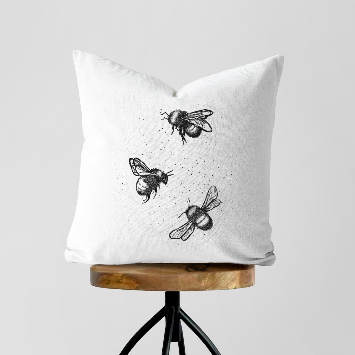 White pillow cover printed with a black ink illustration of three bumble bees flying, on a wooden stool.