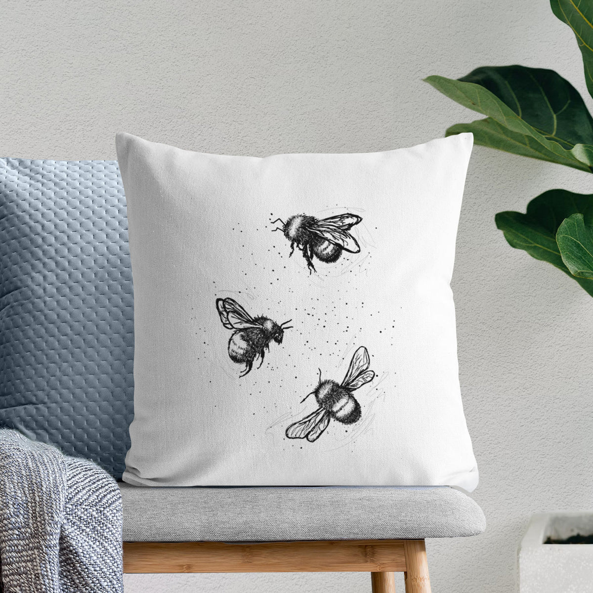 White pillow cover printed with a black ink illustration of three bumble bees flying, on a bench beside plants.