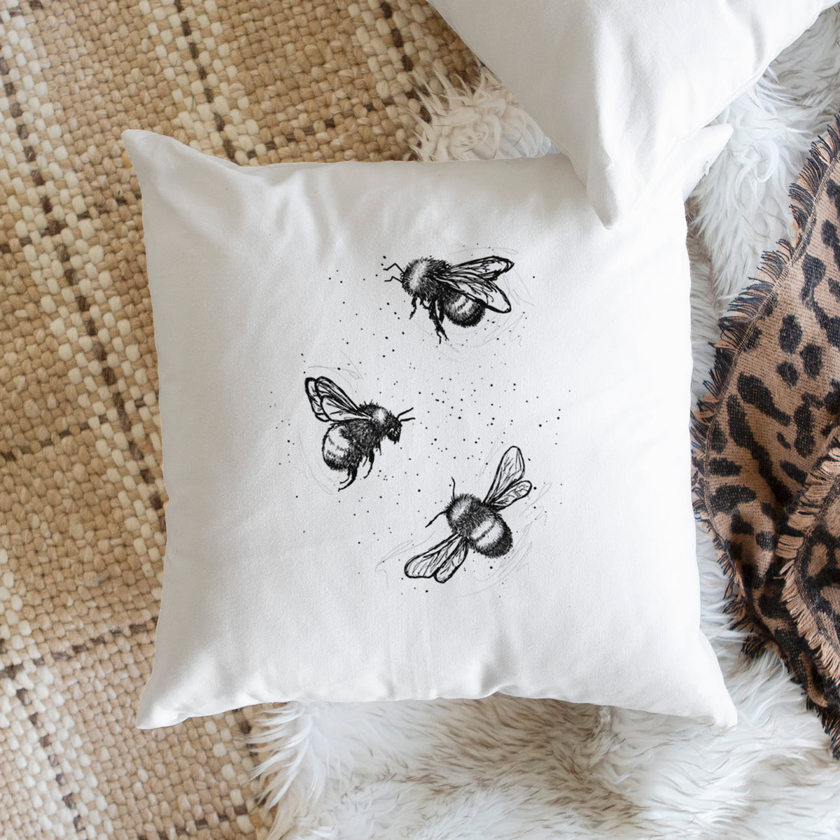 White pillow cover printed with a black ink illustration of three bumble bees flying, on a woven chair.