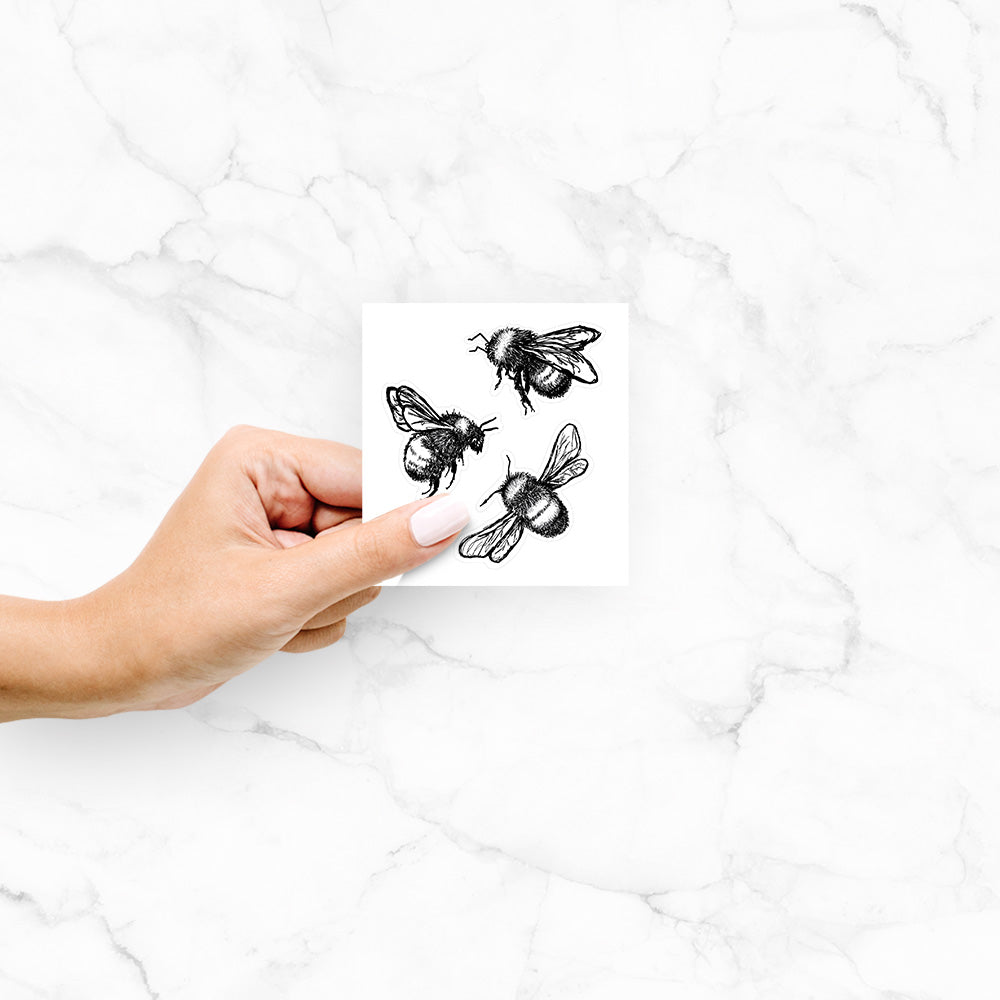 Hand holding a 4 inch vinyl decal of a black ink illustration of three bumble bees flying.