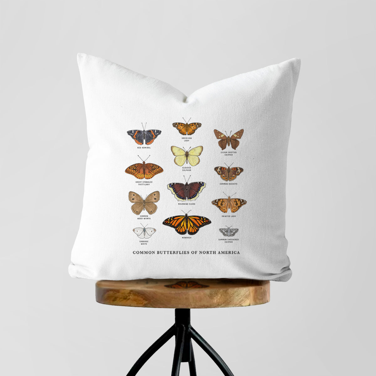 White pillow cover printed with a full-color chart of common butterflies of North America, each labeled with its species, on a wooden stool.