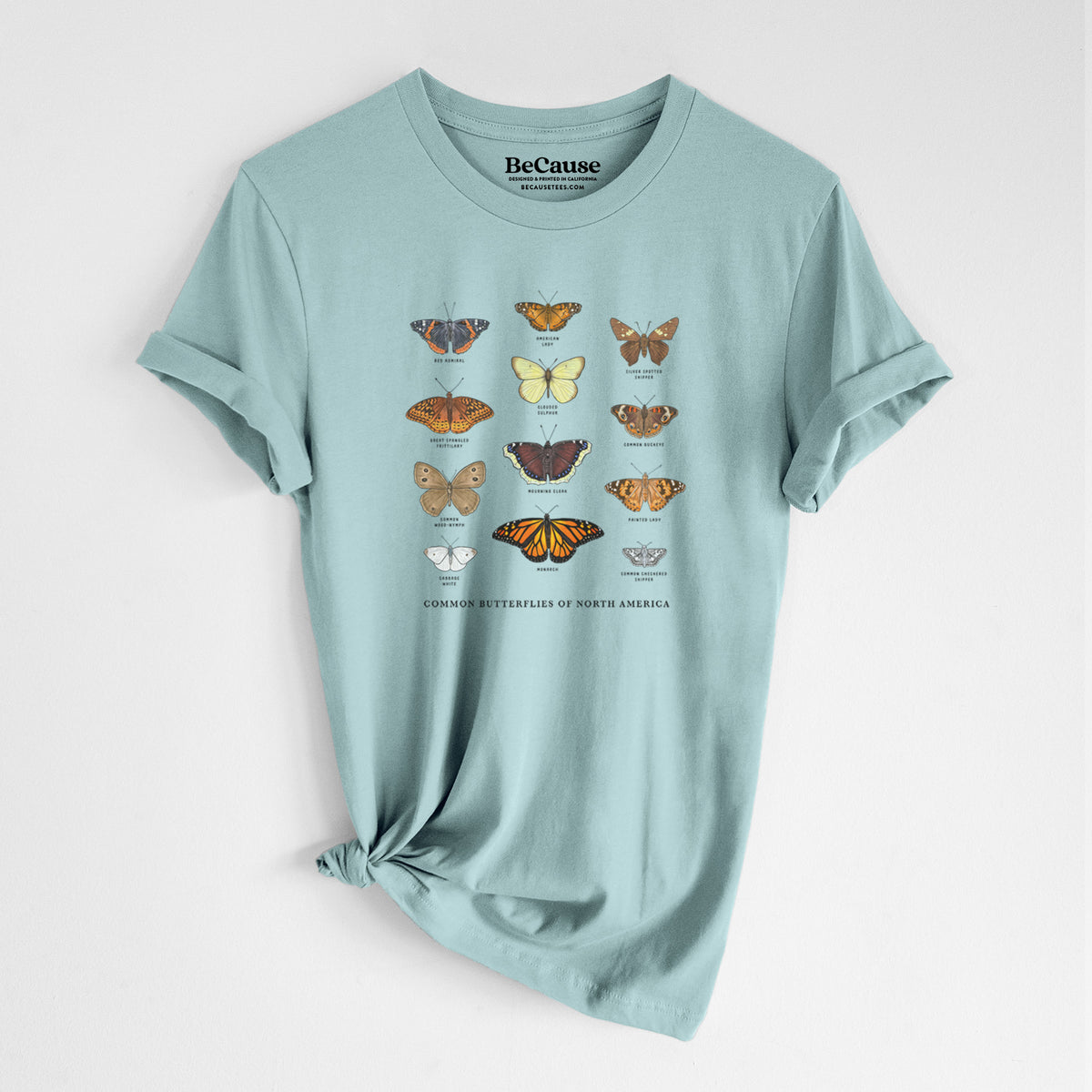 Dusty blue lightweight cotton t-shirt laid flat with the sleeves rolled, printed with a full-color chart of common butterflies of North America, each labeled with its species.