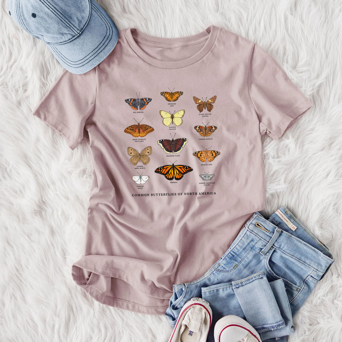 Women's relaxed fit cotton t-shirt printed with a full-color chart of common butterflies of North America, each labeled with its species, styled flat on a white faux fur rug with jeans.