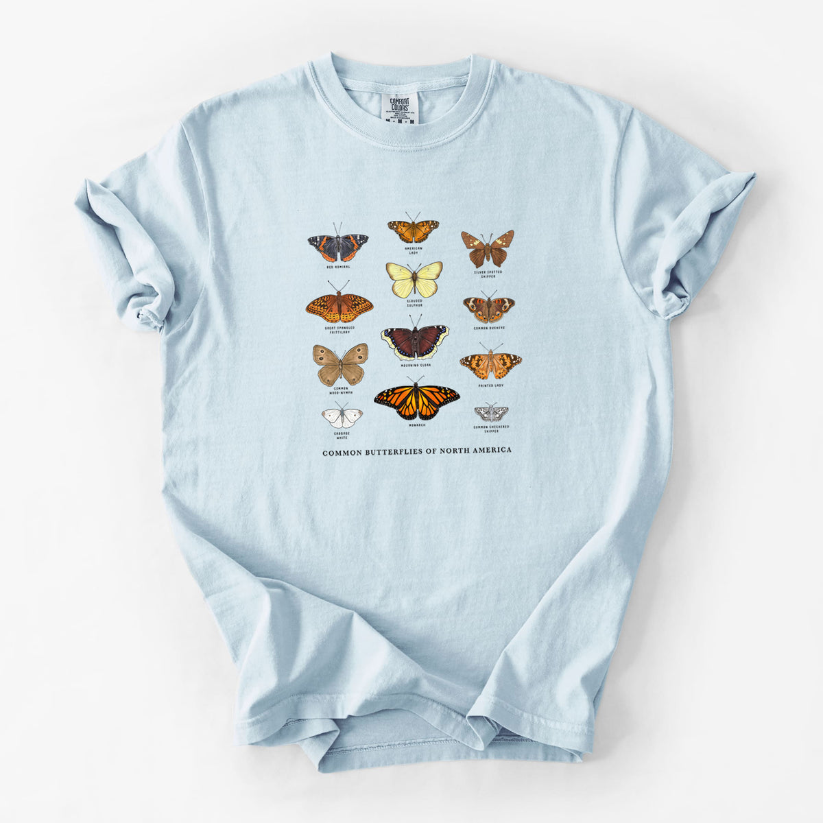 Chambray blue Comfort Colors t-shirt laid flat, printed with a full-color chart of common butterflies of North America, each labeled with its species.