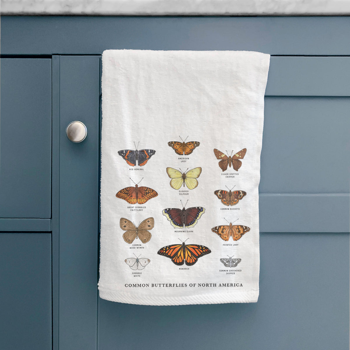 White hand towel printed with a full-color chart of common butterflies of North America, each labeled with its species, hanging on a cabinet door.