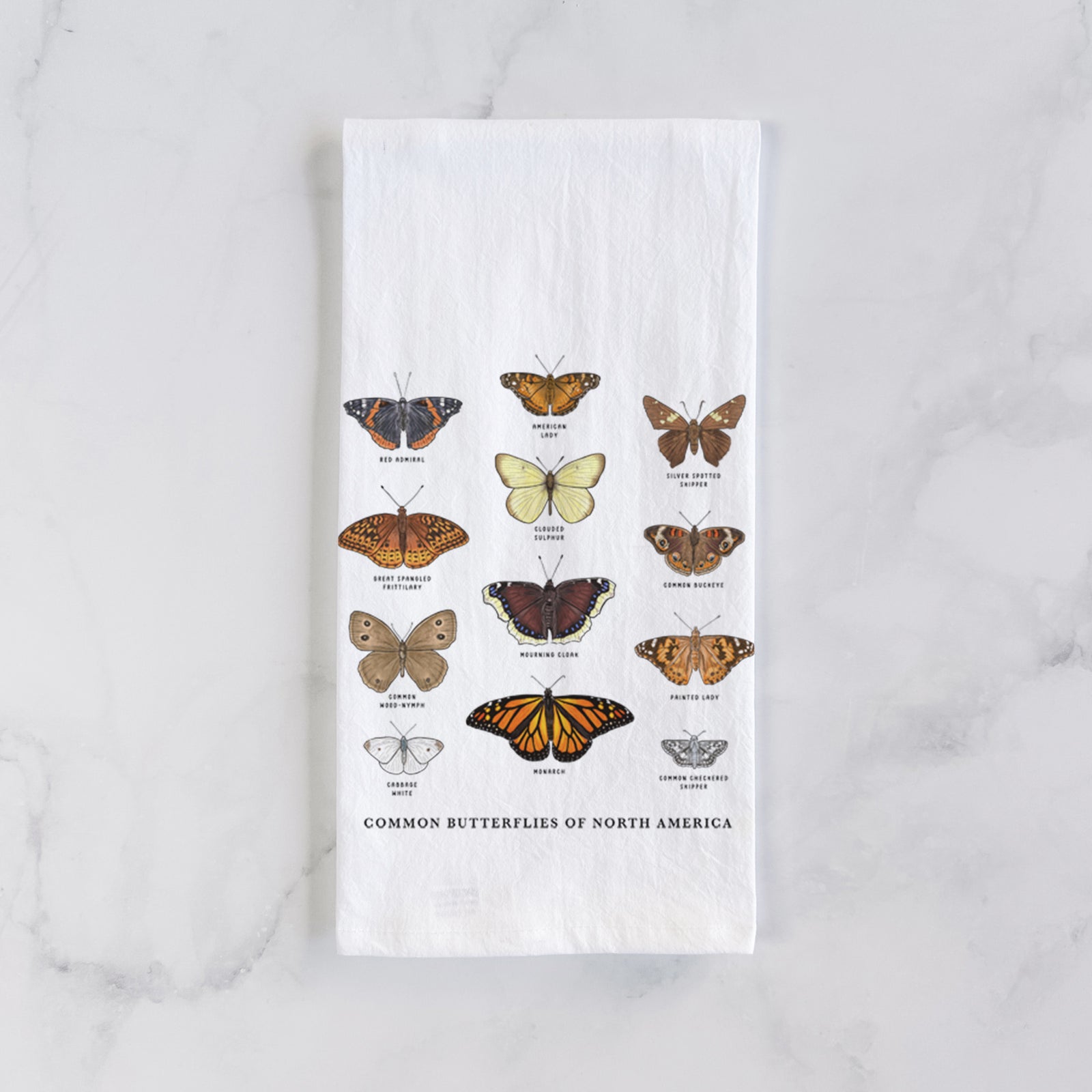 White cotton tea towel printed with a full-color chart of common butterflies of North America, each labeled with its species, on a marble counter beside a round wooden cutting board and greenery.