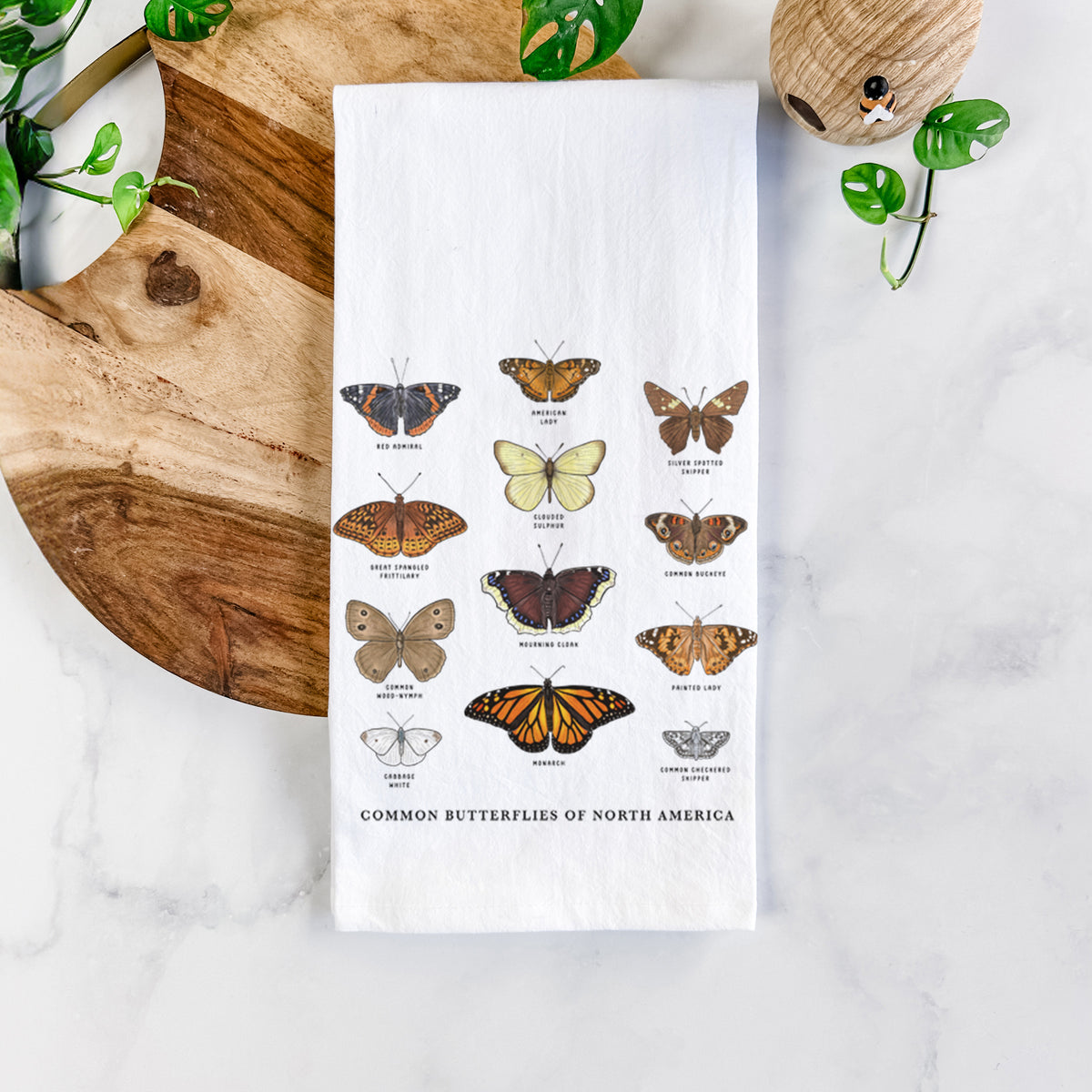 White cotton tea towel printed with a full-color chart of common butterflies of North America, each labeled with its species, on a marble counter beside a round wooden cutting board and greenery.