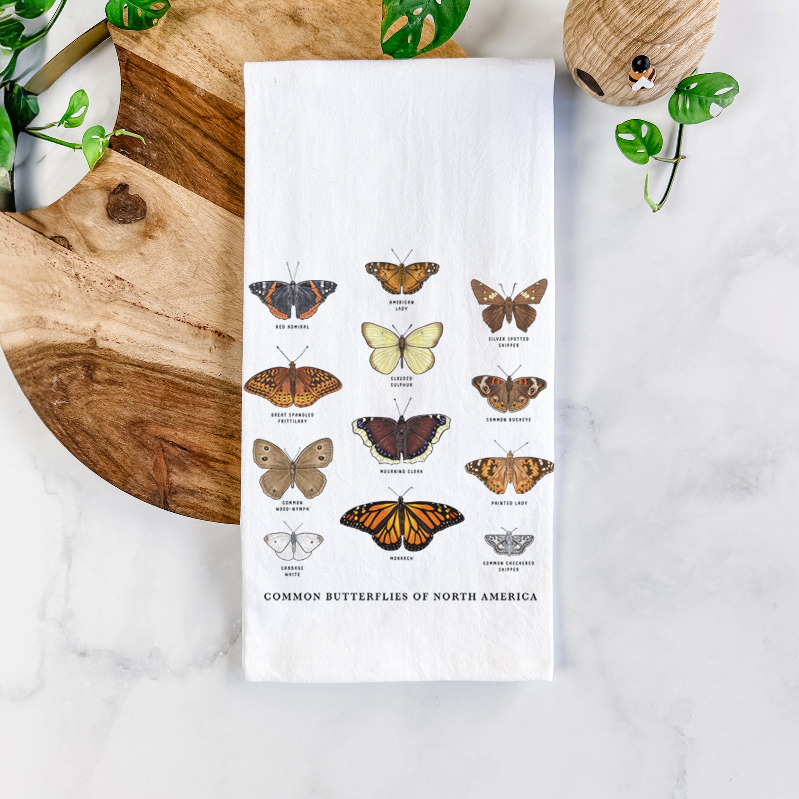 White cotton tea towel printed with a full-color chart of common butterflies of North America, each labeled with its species, on a marble counter beside a round wooden cutting board and greenery.