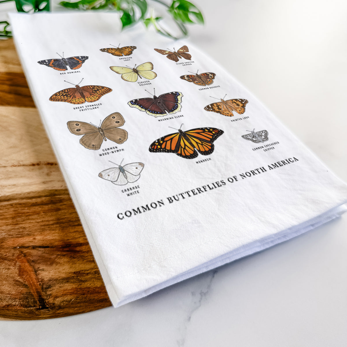 Close-up of a folded white tea towel on a wooden board, showing a full-color chart of common butterflies of North America, each labeled with its species.