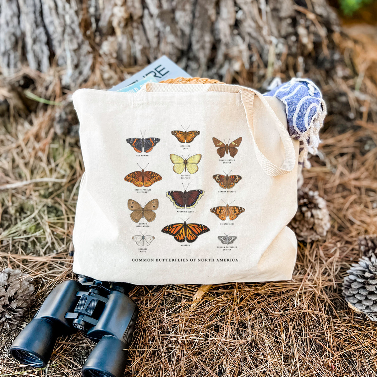 Canvas tote bag printed with a full-color chart of common butterflies of North America, each labeled with its species, resting on dry grass beside a pair of binoculars.