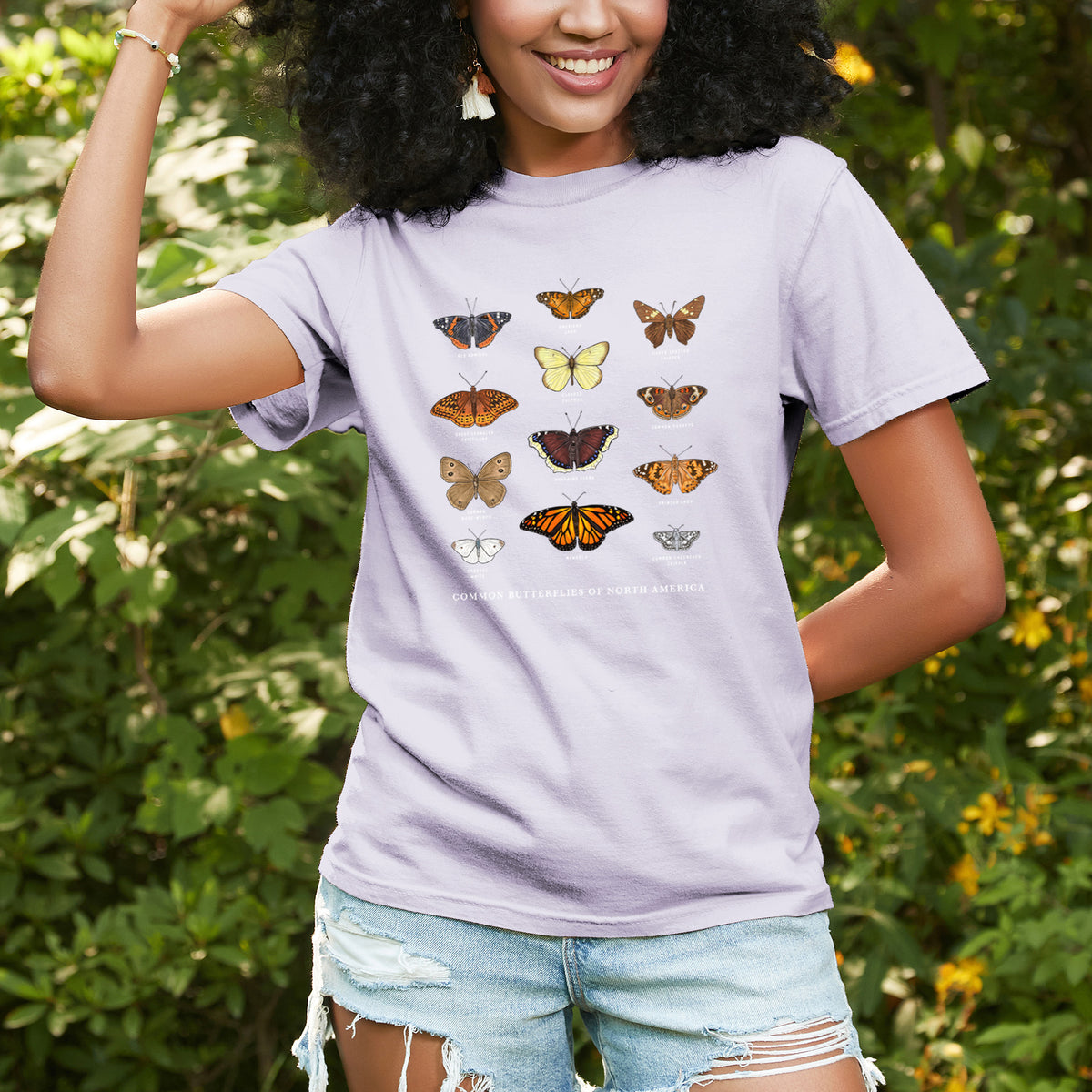 Woman wearing a Comfort Colors t-shirt printed with a full-color chart of common butterflies of North America, each labeled with its species, outdoors.