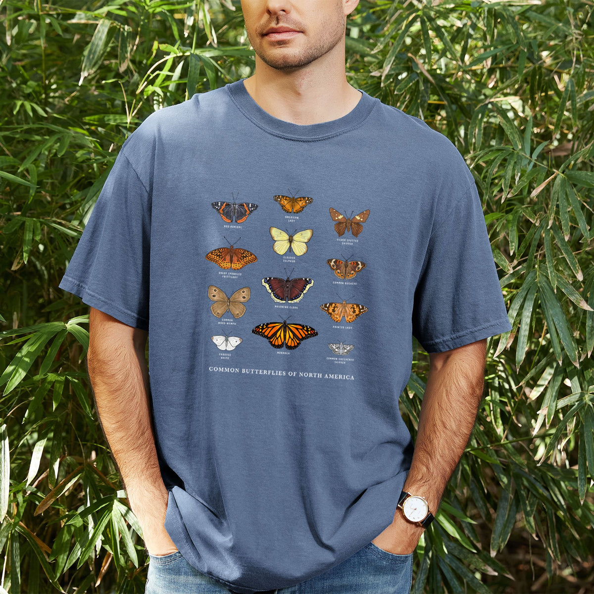 Man wearing a Comfort Colors t-shirt printed with a full-color chart of common butterflies of North America, each labeled with its species, outdoors.