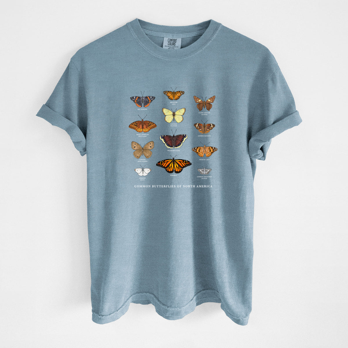 Steel blue Comfort Colors t-shirt laid flat, printed with a full-color chart of common butterflies of North America, each labeled with its species.