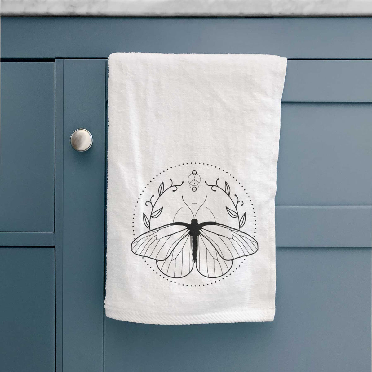 White hand towel printed with a black ink butterfly inside a circle, framed by moon phases and leaves, hanging on a cabinet door.