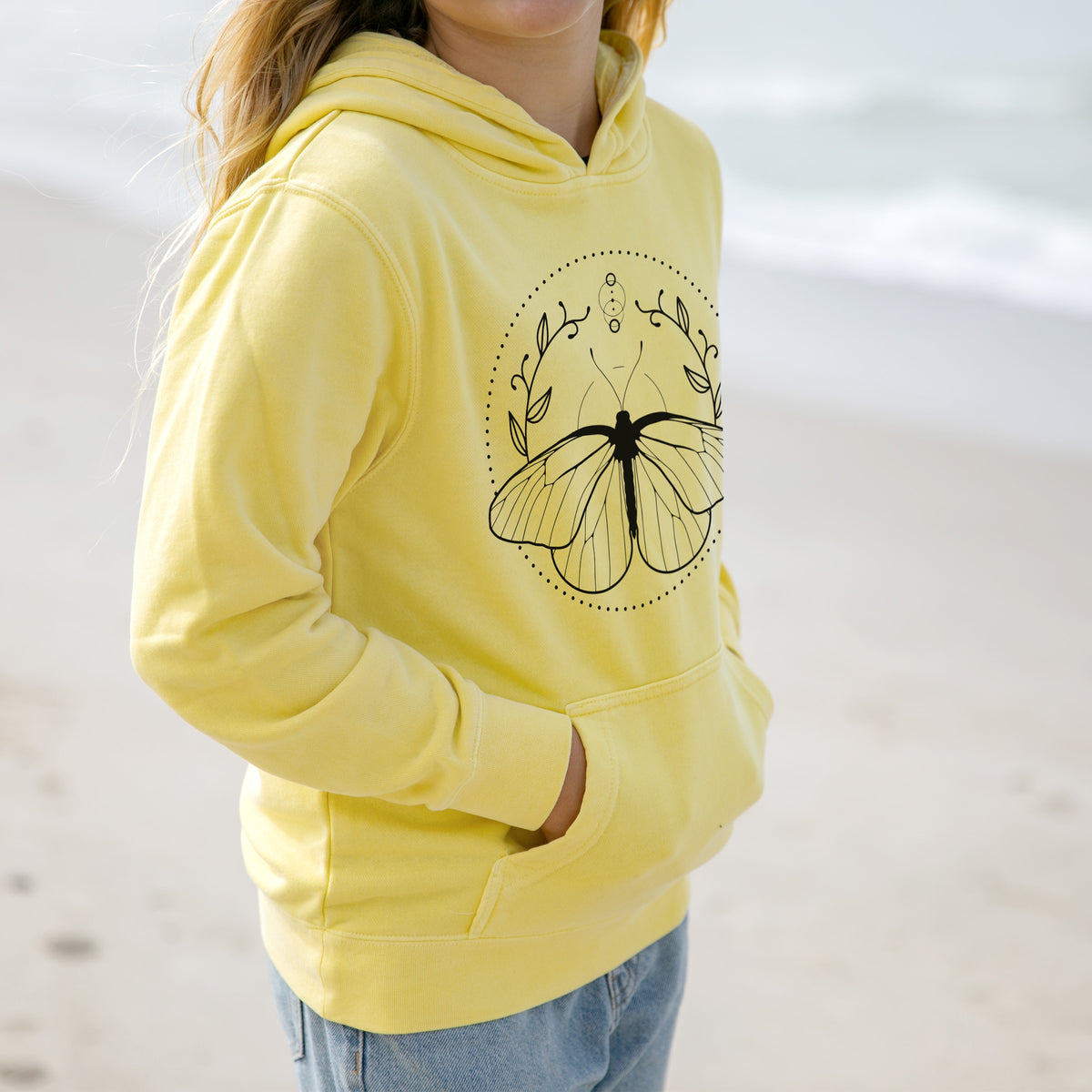 Girl wearing a youth pigment dyed hoodie printed with a black ink butterfly inside a circle, framed by moon phases and leaves.