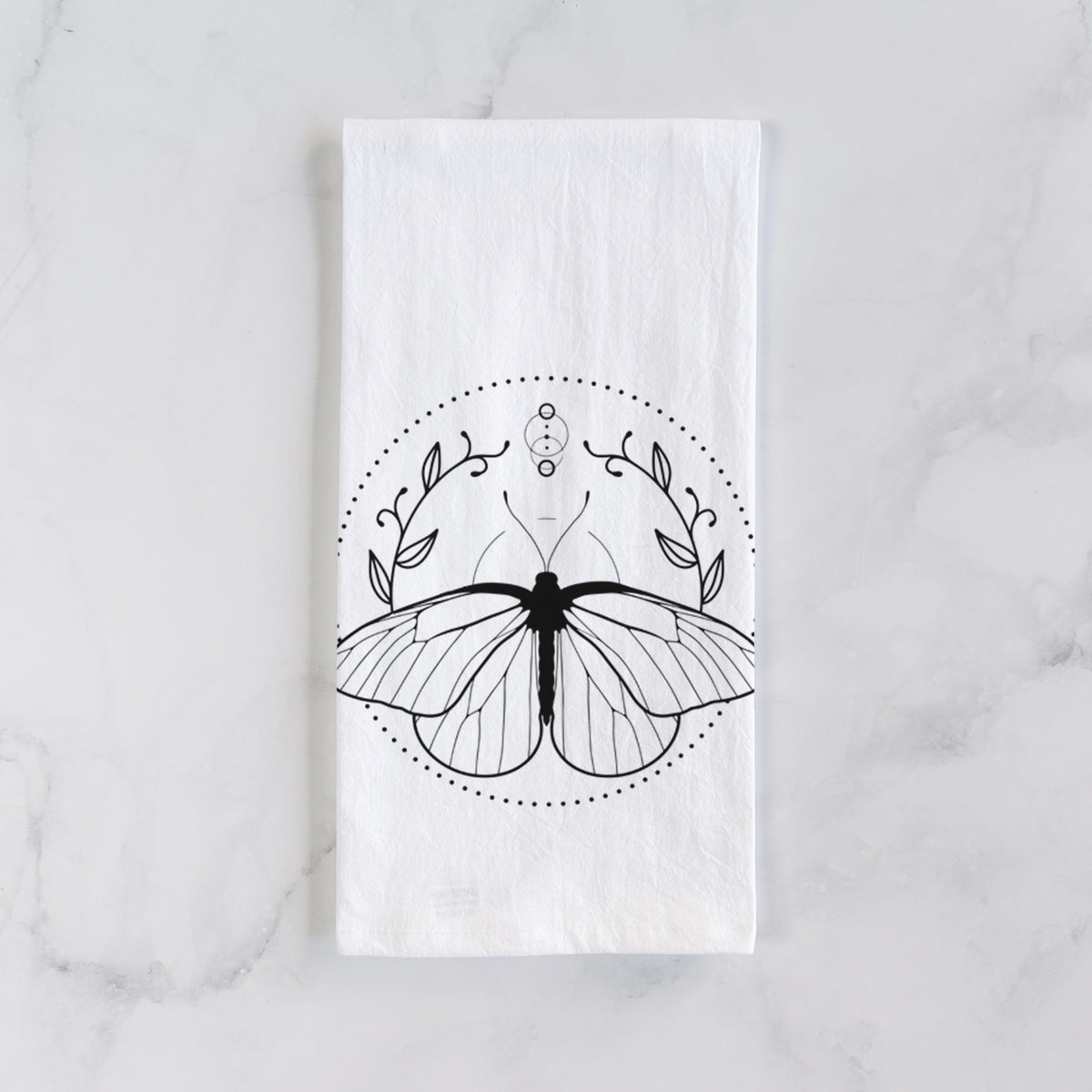 White cotton tea towel laid flat on marble, printed with a black ink butterfly inside a circle, framed by moon phases and leaves.