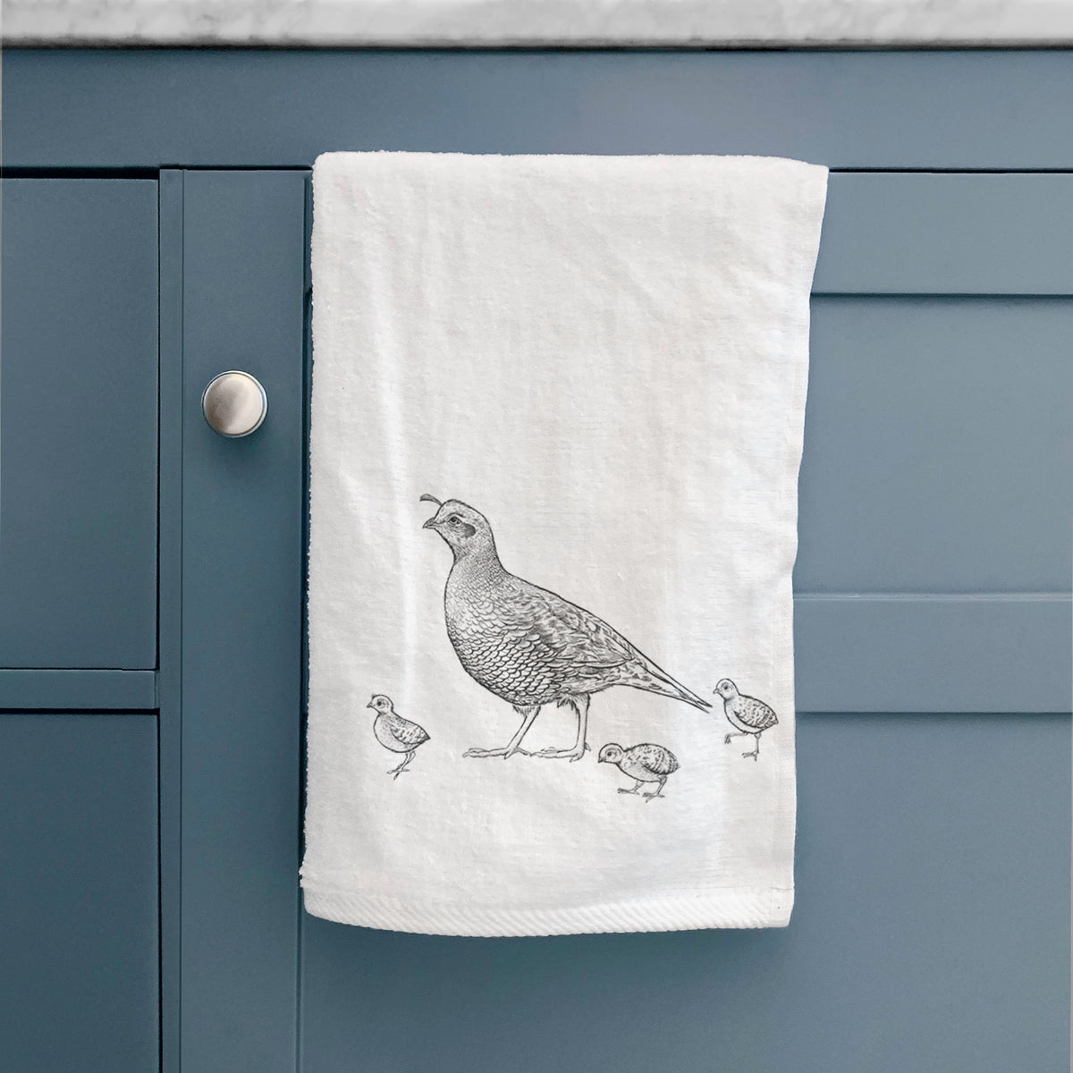 White hand towel printed with a black ink illustration of a California quail with its chicks, hanging on a cabinet door.