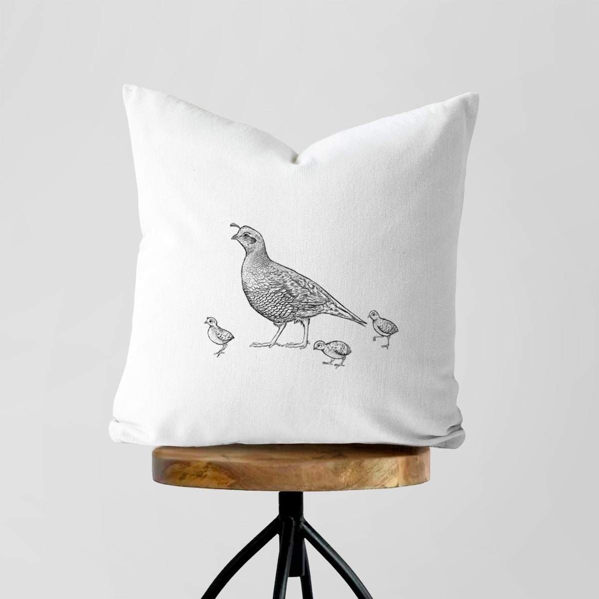 White pillow cover printed with a black ink illustration of a California quail with its chicks, on a wooden stool.