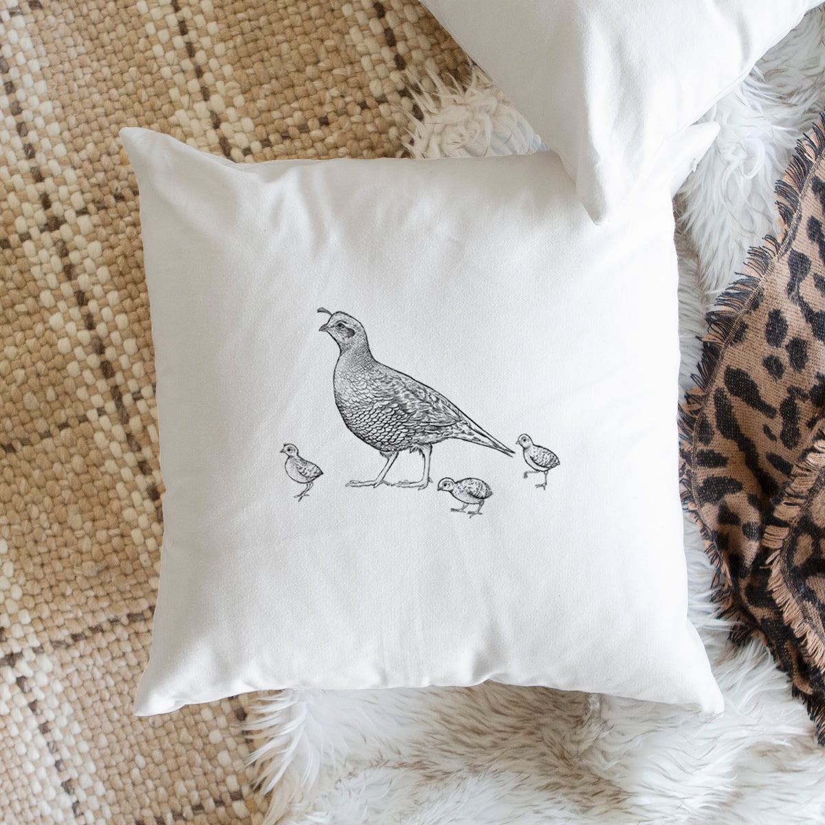 White pillow cover printed with a black ink illustration of a California quail with its chicks, on a woven chair.