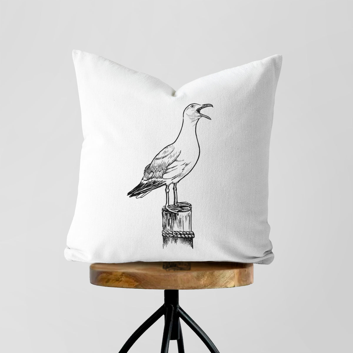 White pillow cover printed with a black ink illustration of a California gull calling from a wooden post, on a wooden stool.