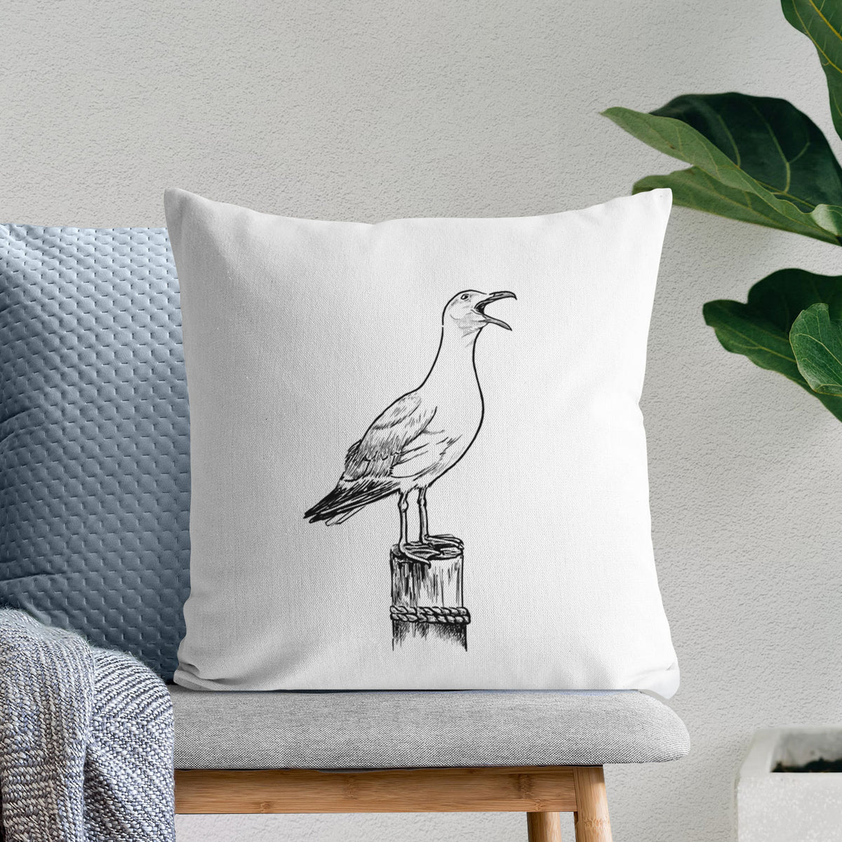 White pillow cover printed with a black ink illustration of a California gull calling from a wooden post, on a bench beside plants.