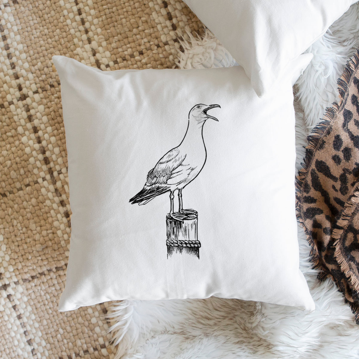 White pillow cover printed with a black ink illustration of a California gull calling from a wooden post, on a woven chair.