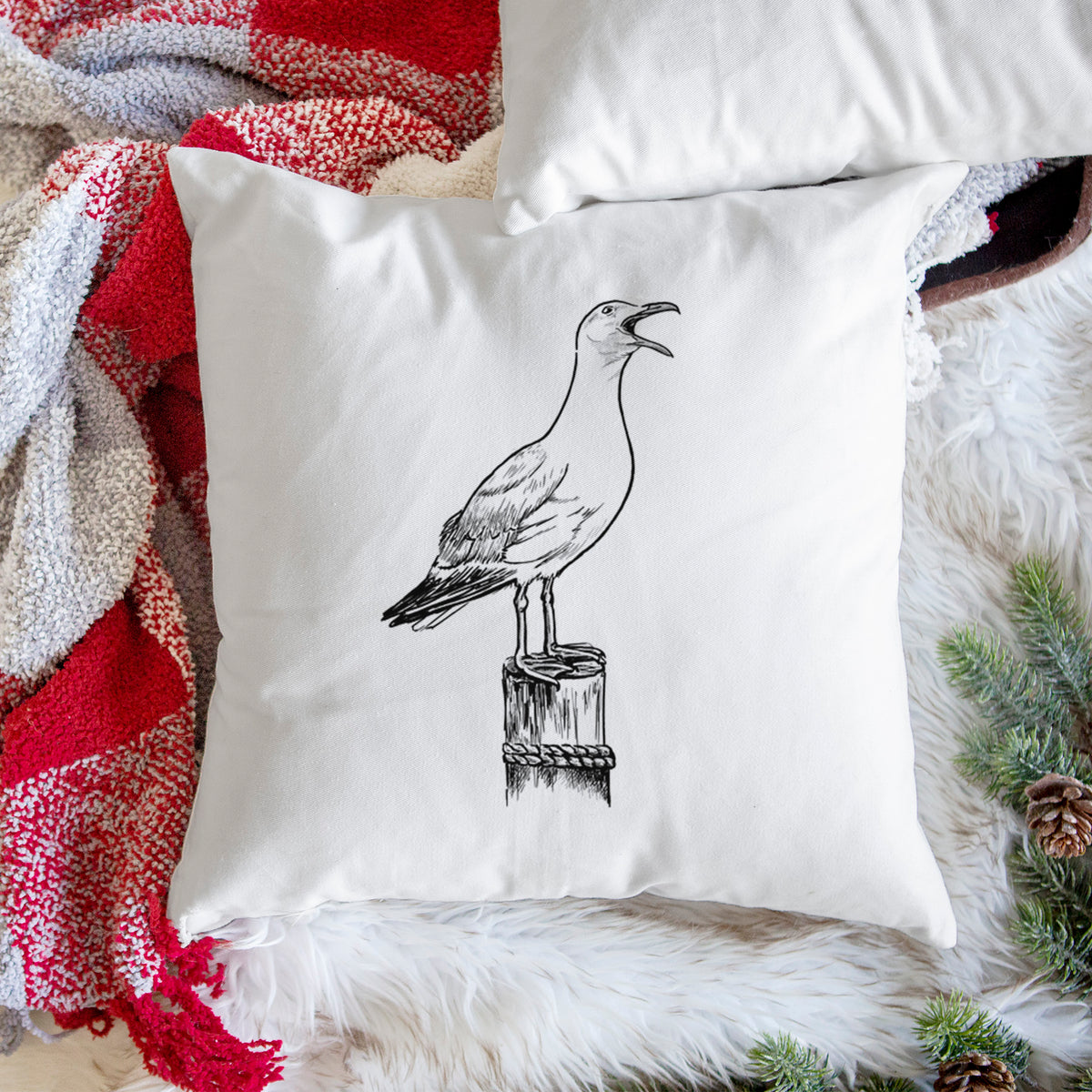 White pillow cover printed with a black ink illustration of a California gull calling from a wooden post, on a woven chair.