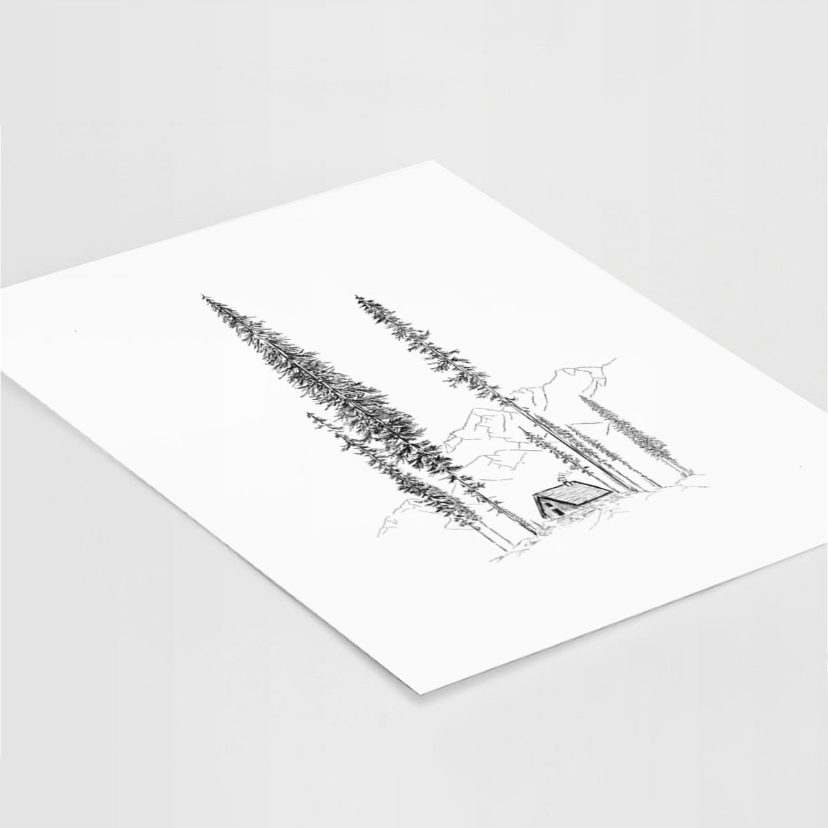 Art print of a black ink illustration of a small cabin nestled beneath towering pine trees, lying at an angle on a white surface.
