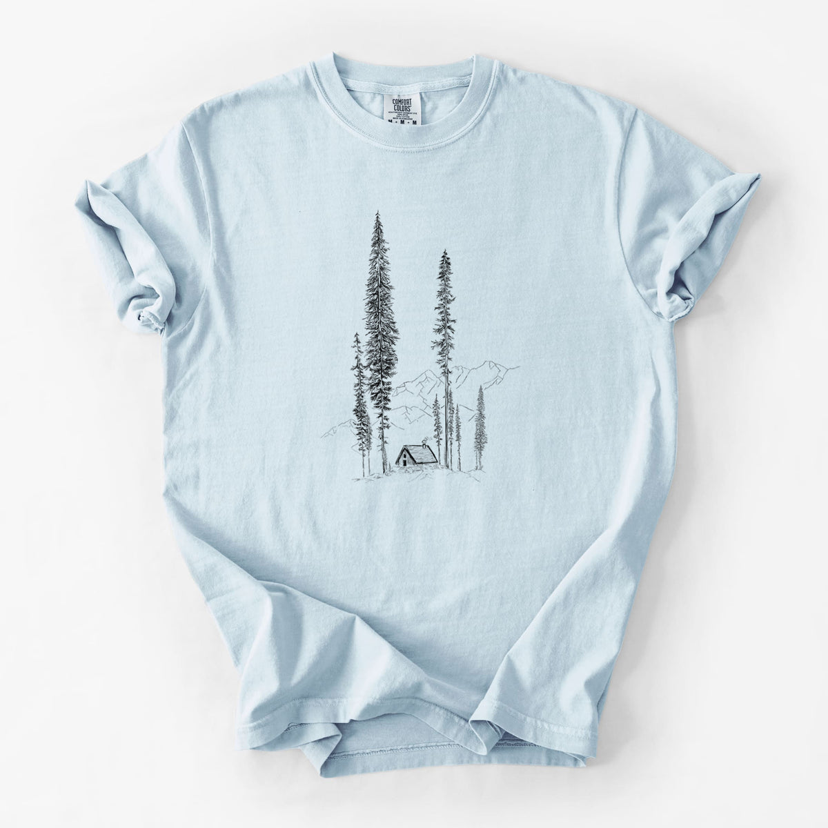 Chambray blue Comfort Colors t-shirt laid flat, printed with a black ink illustration of a small cabin nestled beneath towering pine trees.