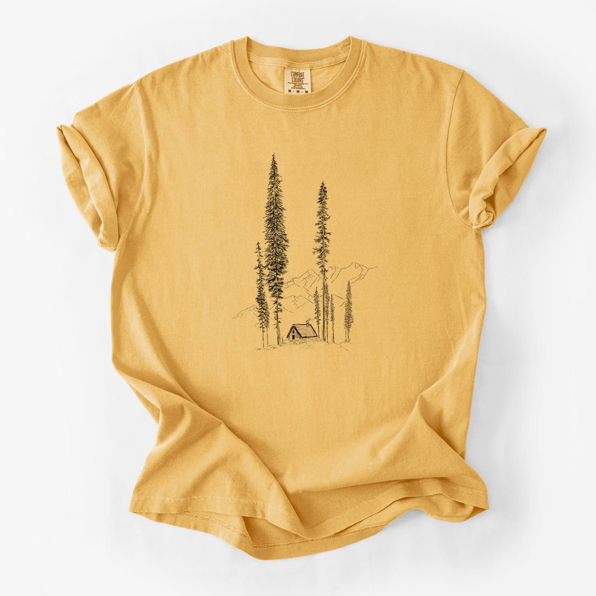 Golden wheat yellow Comfort Colors t-shirt laid flat, printed with a black ink illustration of a small cabin nestled beneath towering pine trees.