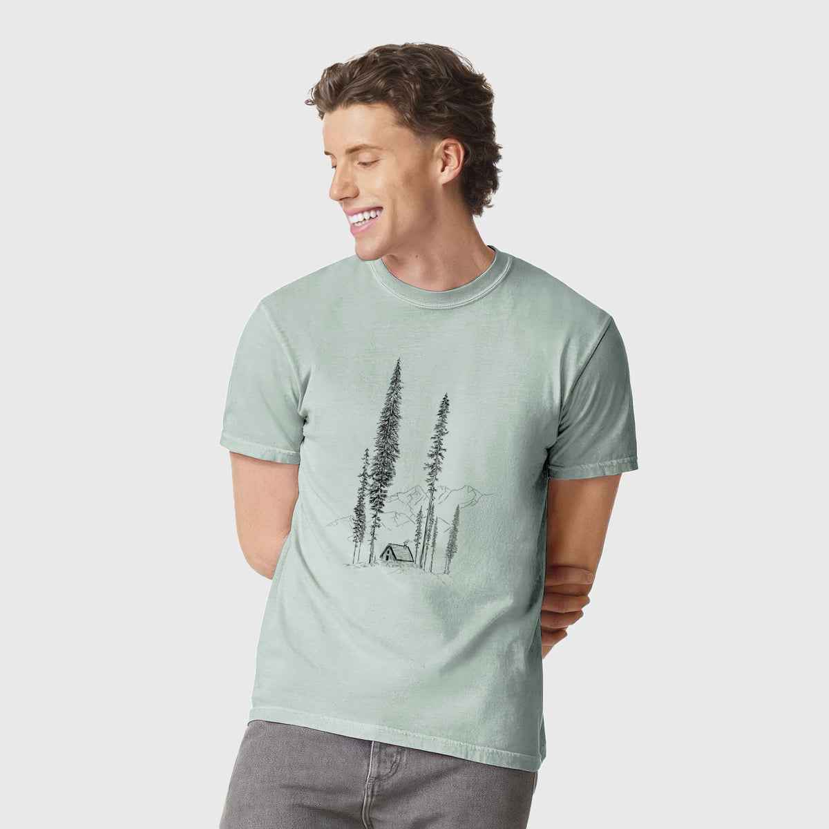 Man wearing a Comfort Colors t-shirt printed with a black ink illustration of a small cabin nestled beneath towering pine trees.