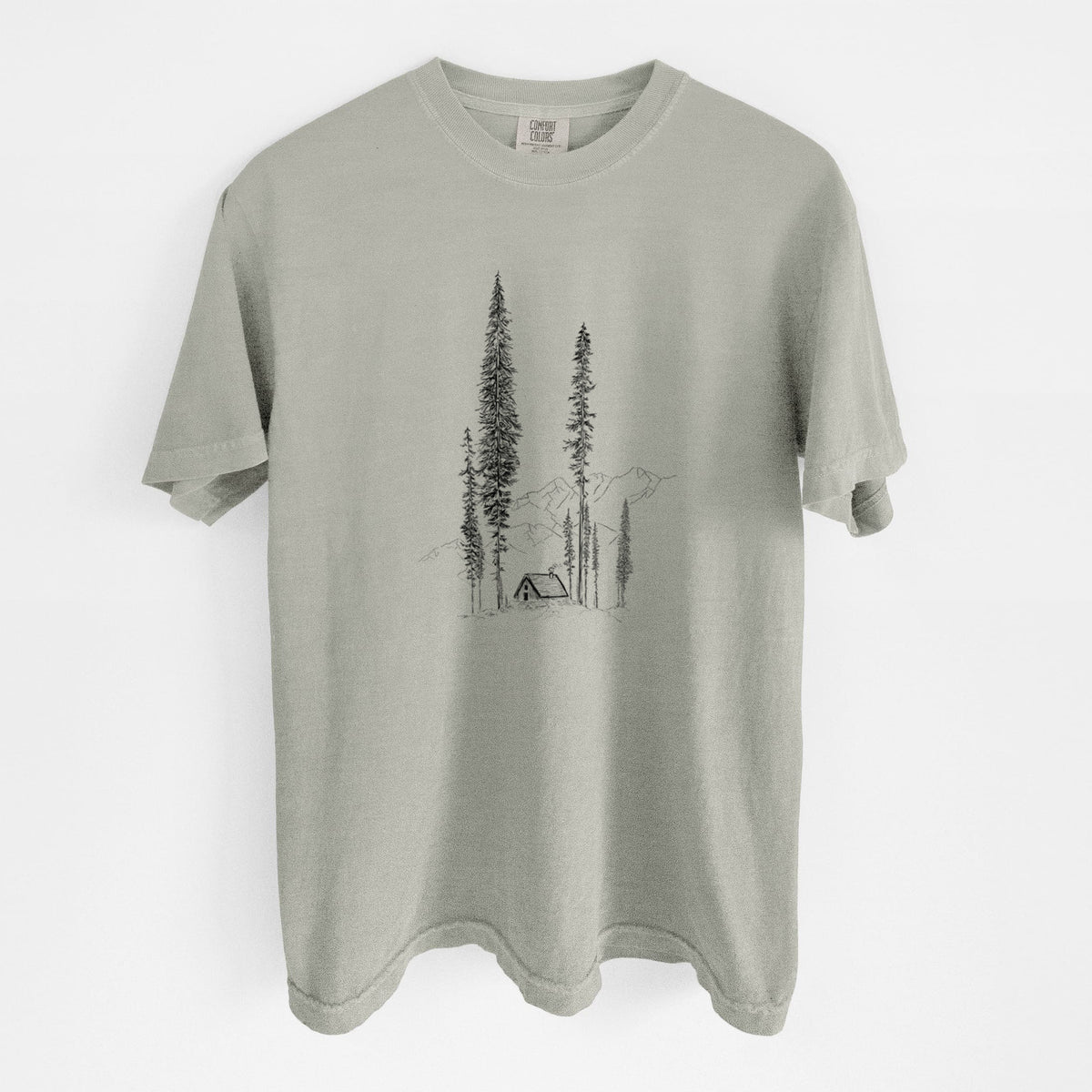 Sandstone beige Comfort Colors t-shirt laid flat, printed with a black ink illustration of a small cabin nestled beneath towering pine trees.