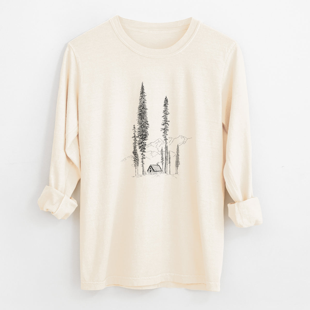 Ivory Comfort Colors long sleeve shirt laid flat with the cuffs rolled, printed with a black ink illustration of a small cabin nestled beneath towering pine trees.