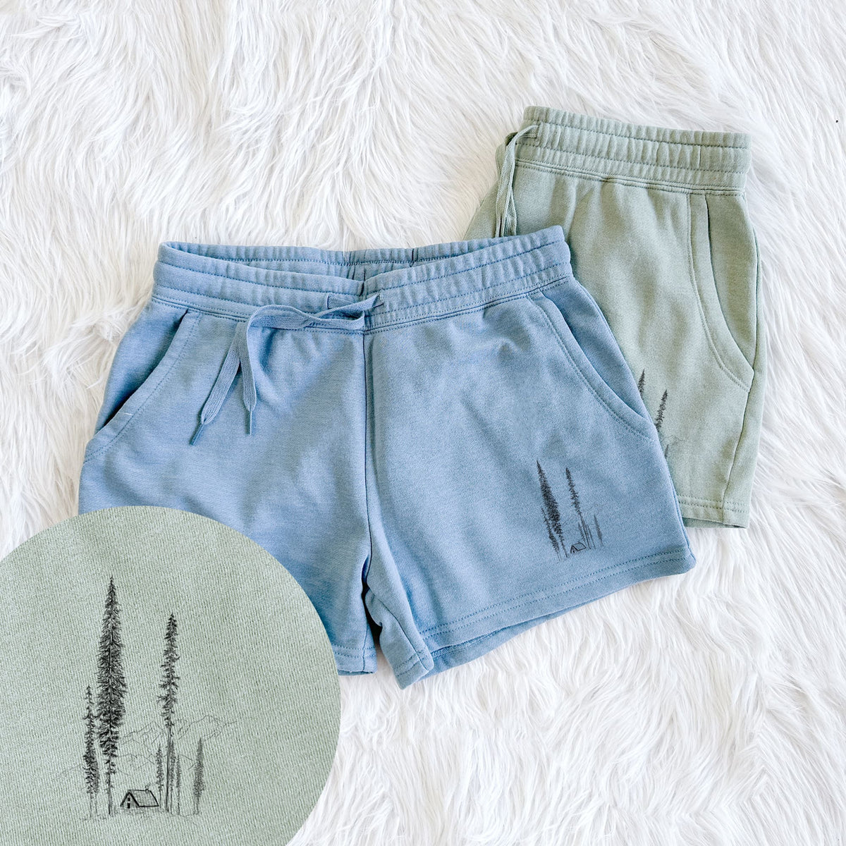 Two pairs of Cali Wave shorts in different colors printed with a black ink illustration of a small cabin nestled beneath towering pine trees, with a close-up of the print.