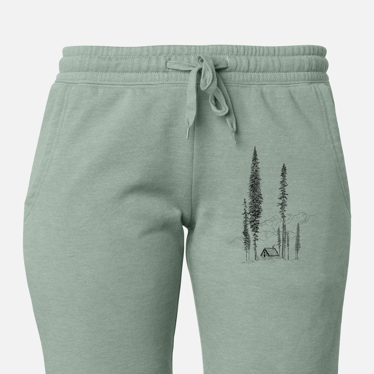 Front of sage green Cali Wave sweatpants, printed with a black ink illustration of a small cabin nestled beneath towering pine trees on the leg.