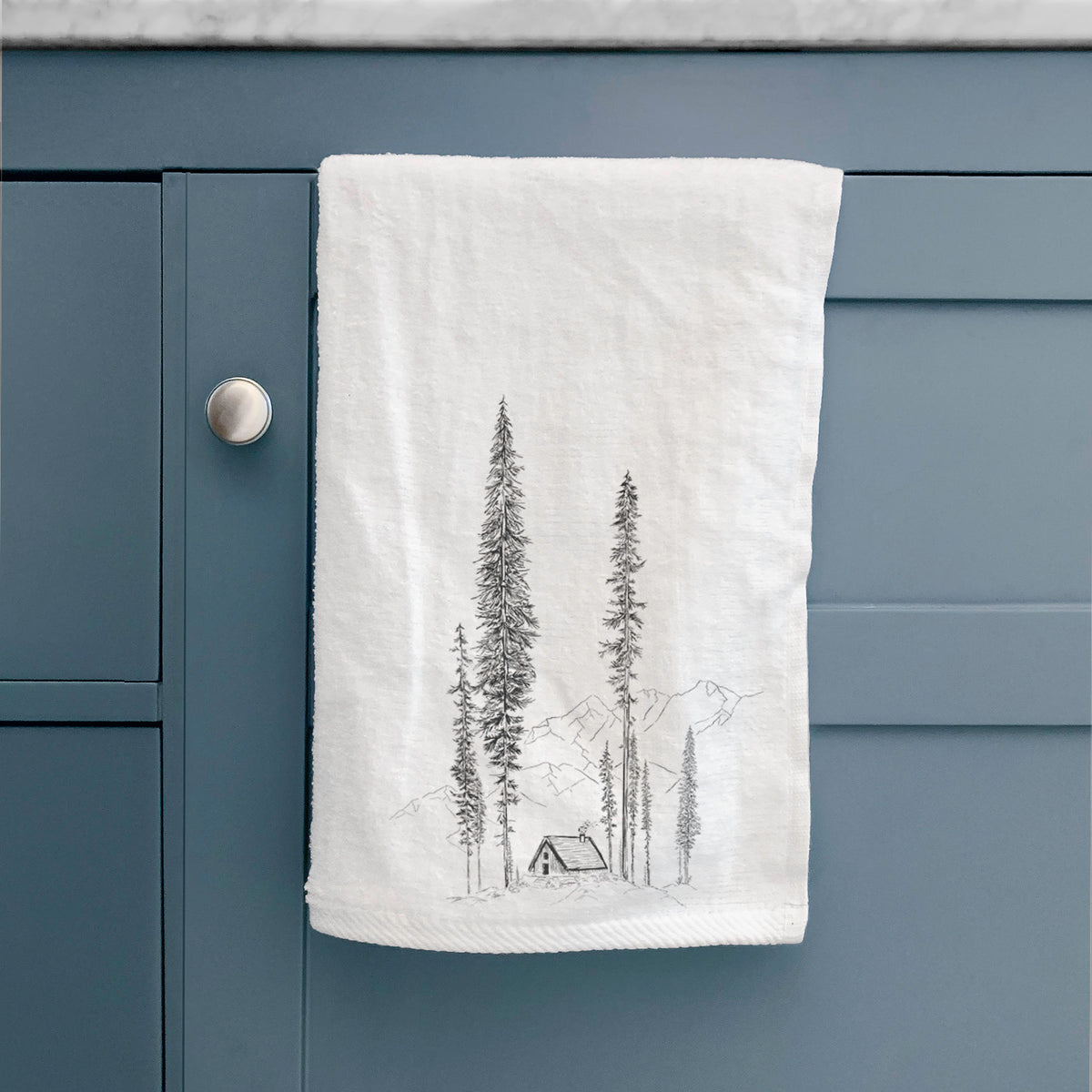 White hand towel printed with a black ink illustration of a small cabin nestled beneath towering pine trees, hanging on a cabinet door.