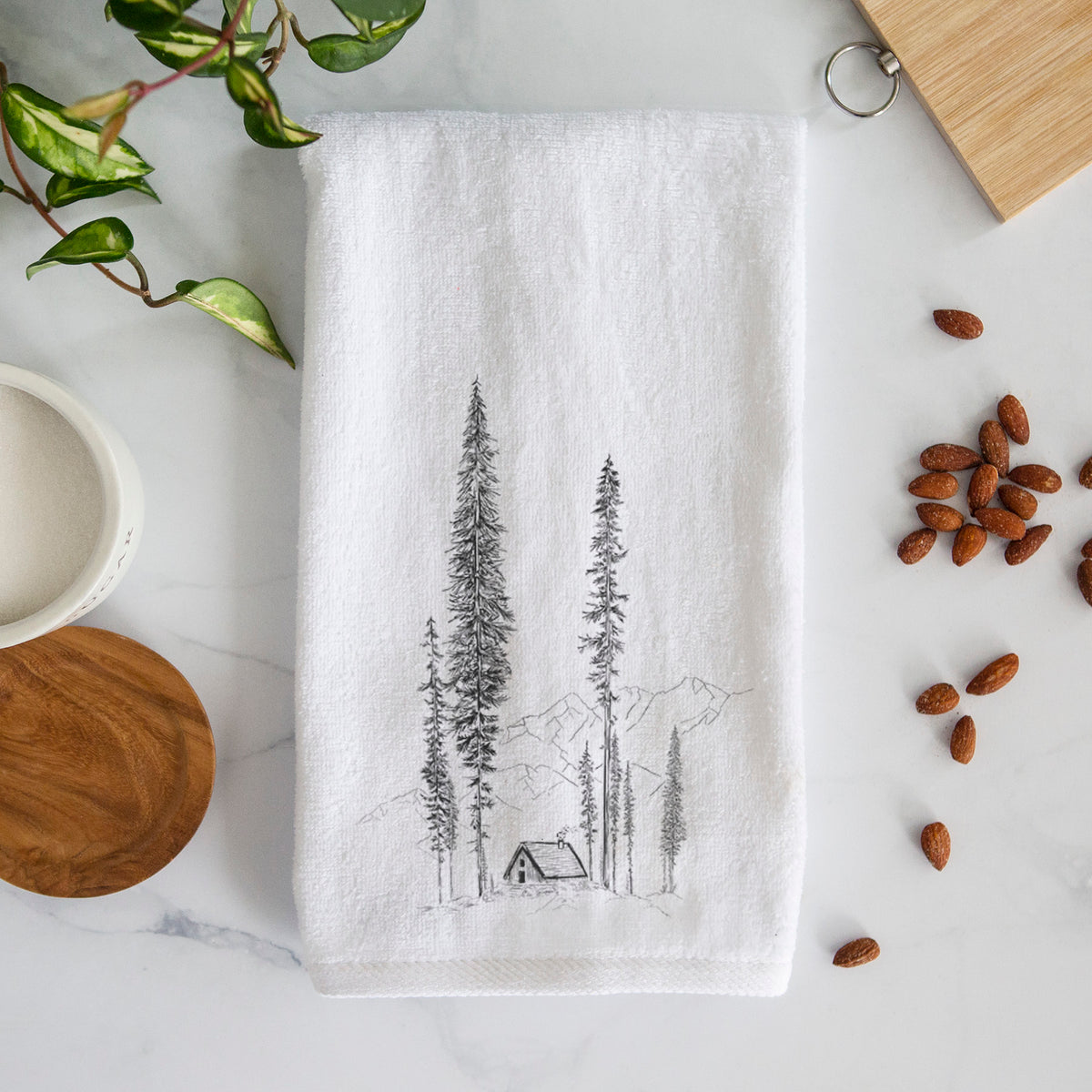 White hand towel printed with a black ink illustration of a small cabin nestled beneath towering pine trees.