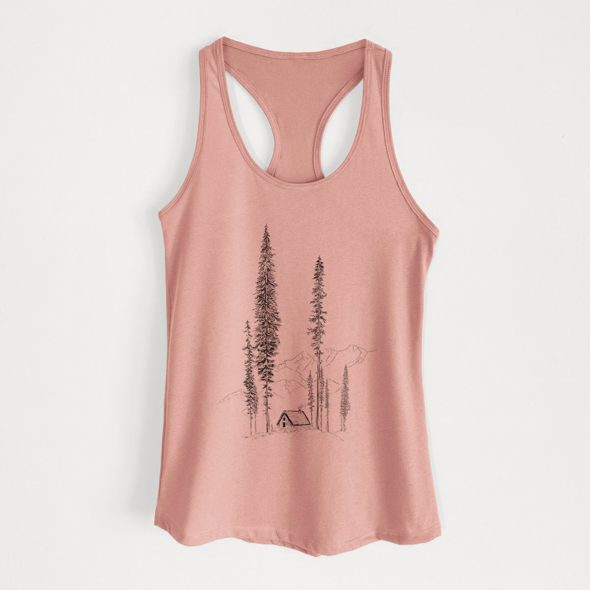 Desert pink women's racerback tank top laid flat, printed with a black ink illustration of a small cabin nestled beneath towering pine trees.