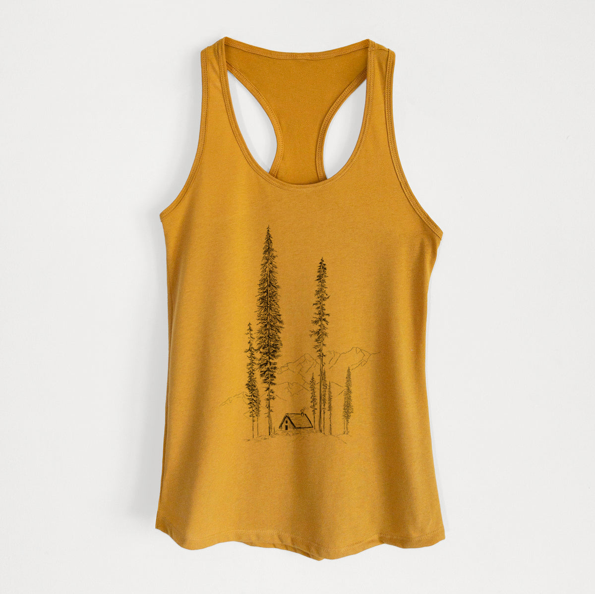 Gold women's racerback tank top laid flat, printed with a black ink illustration of a small cabin nestled beneath towering pine trees.