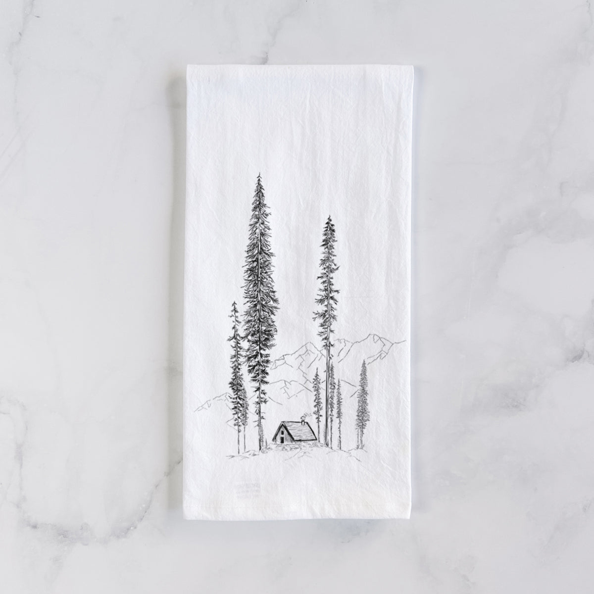 White cotton tea towel laid flat on marble, printed with a black ink illustration of a small cabin nestled beneath towering pine trees.