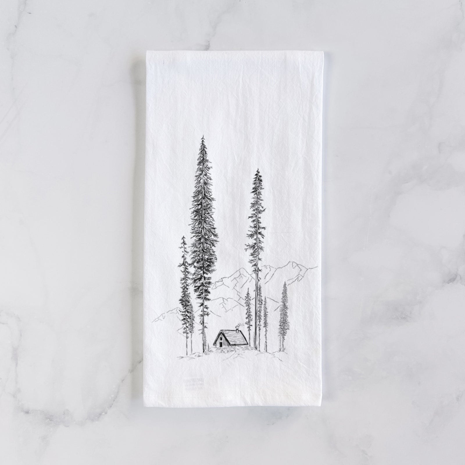 White cotton tea towel printed with a black ink illustration of a small cabin nestled beneath towering pine trees, on a marble counter beside a round wooden cutting board and greenery.