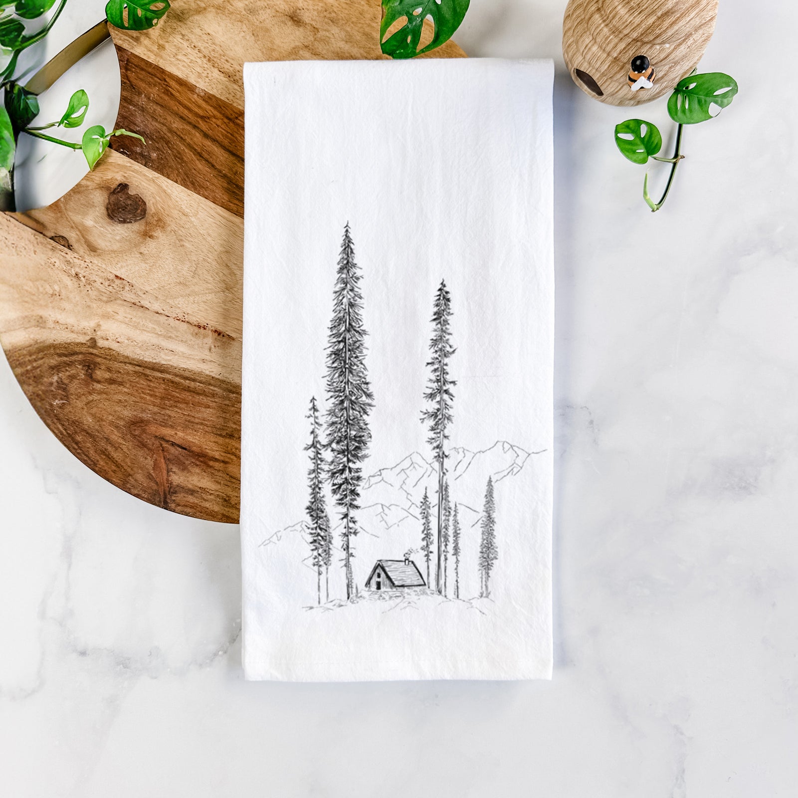 White cotton tea towel printed with a black ink illustration of a small cabin nestled beneath towering pine trees, on a marble counter beside a round wooden cutting board and greenery.