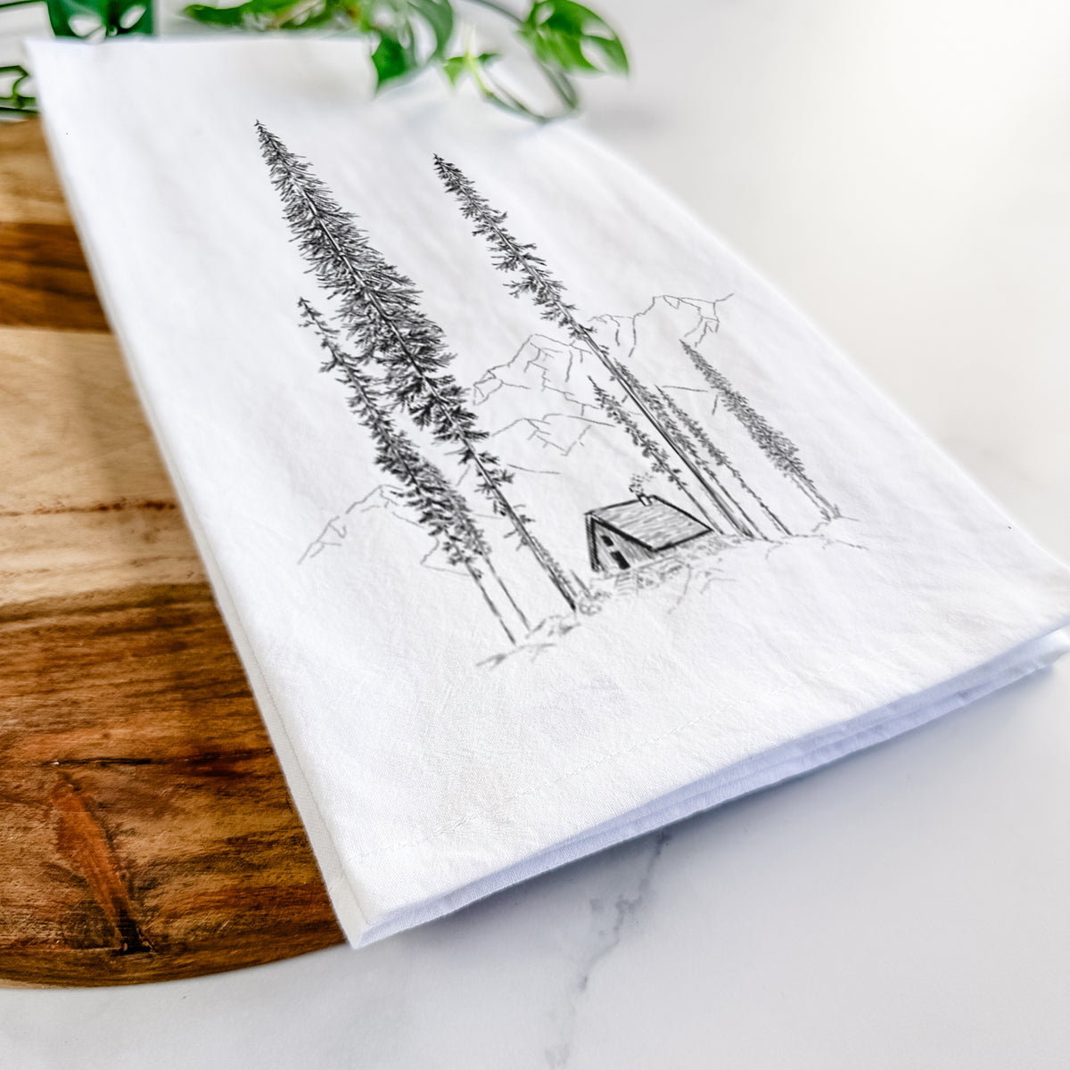 Close-up of a folded white tea towel on a wooden board, showing a black ink illustration of a small cabin nestled beneath towering pine trees.