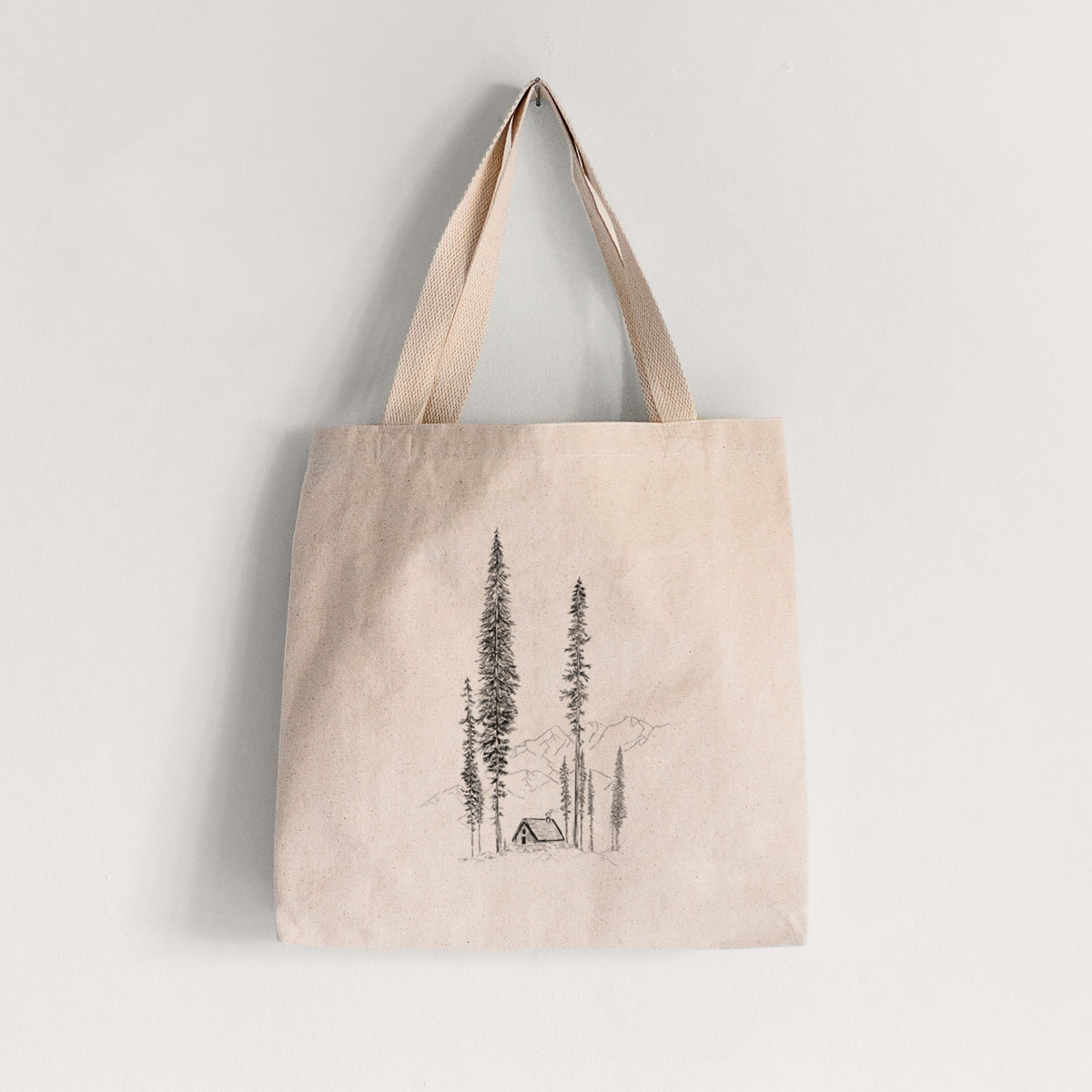 Natural canvas tote bag printed with a black ink illustration of a small cabin nestled beneath towering pine trees.