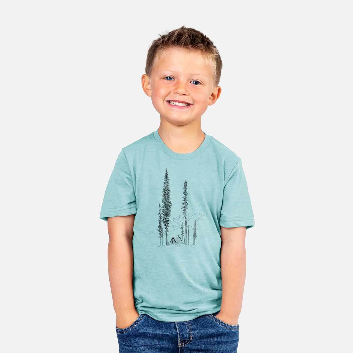 Smiling boy wearing a youth t-shirt printed with a black ink illustration of a small cabin nestled beneath towering pine trees.