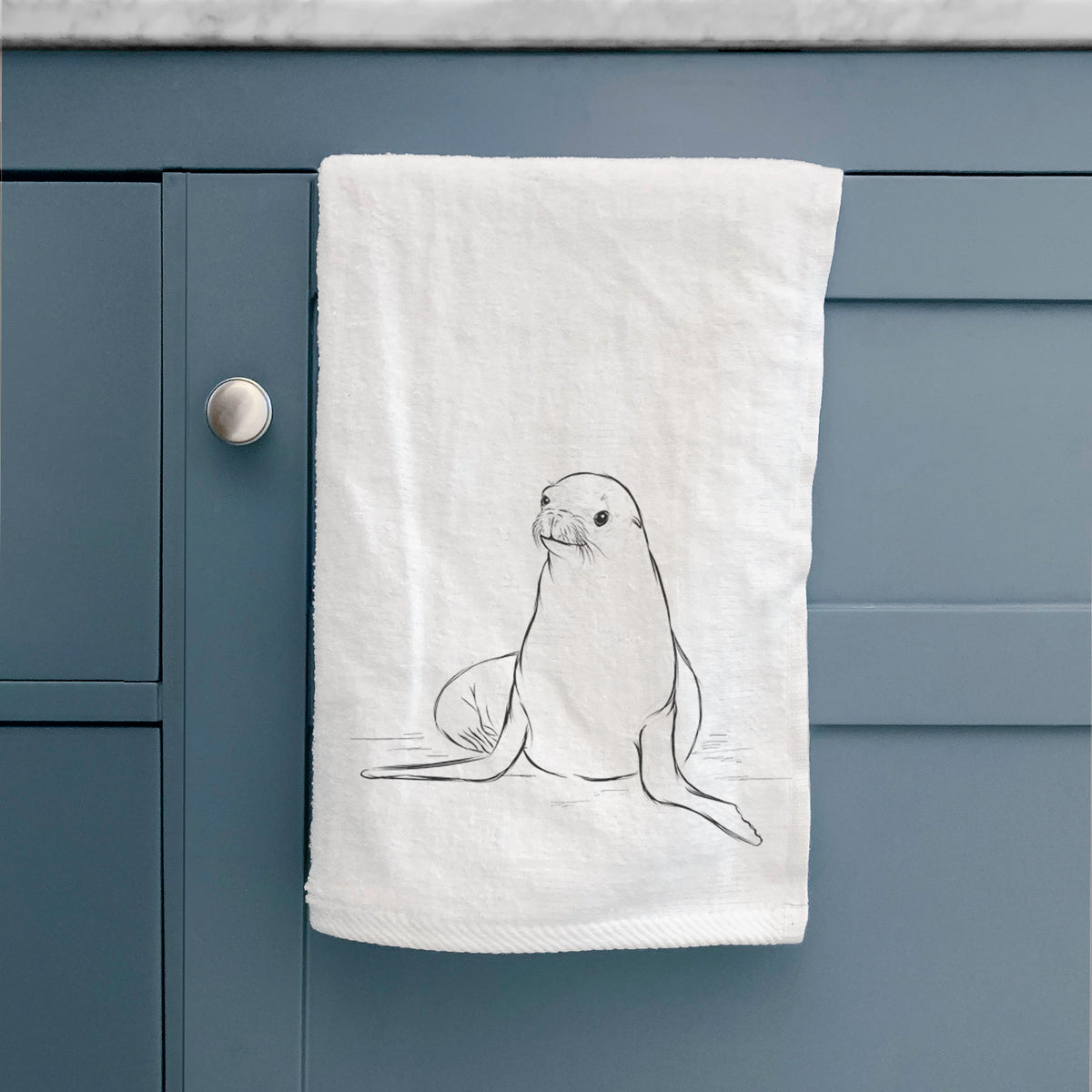 White hand towel printed with a black ink illustration of a California sea lion sitting upright, hanging on a cabinet door.