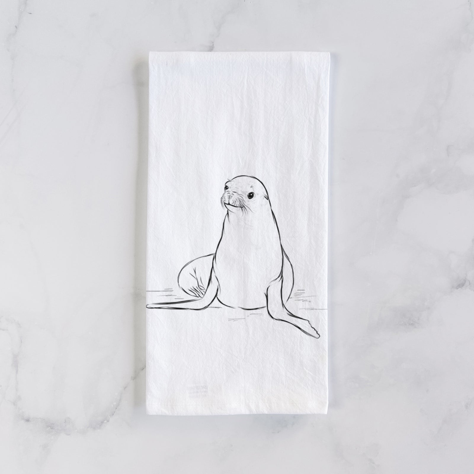 White cotton tea towel printed with a black ink illustration of a California sea lion sitting upright, on a marble counter beside a round wooden cutting board and greenery.